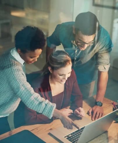 Three people gather around a laptop at a desk, collaborating and smiling as they look at the screen. One person is seated typing, while the other two stand and point at the laptop. The setting appears to be an office.