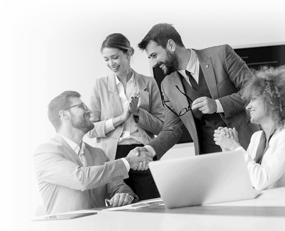 Four business professionals in suits are gathered around a desk. Two are shaking hands and smiling, while the other two stand and sit nearby, clapping and appearing pleased. A laptop is on the desk.